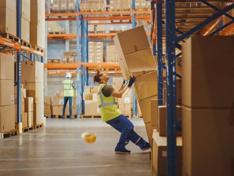 Shot of a Warehouse Worker Has Work Related Accident. He is Falling Down BeforeTrying to Pick Up Heavy Cardboard Box from the Shelf. Hard Injury at Work.